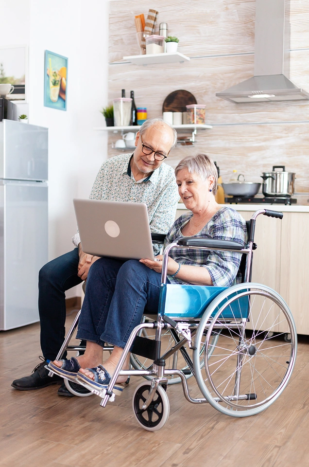 Senior couple at home looking at a laptop, with the man seated in a manual wheelchair to support independent living. Senior couple at home looking at a laptop, with the man seated in a manual wheelchair to support independent living.