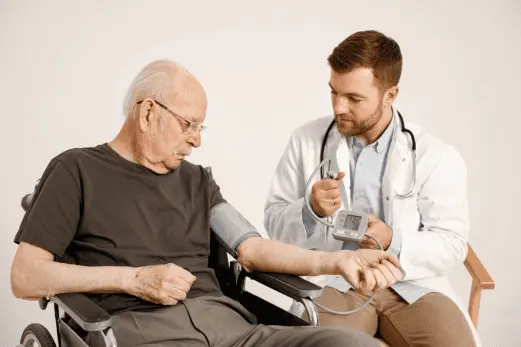A doctor taking the blood pressure of an elderly male patient seated in a wheelchair, using professional diagnostic medical equipment.